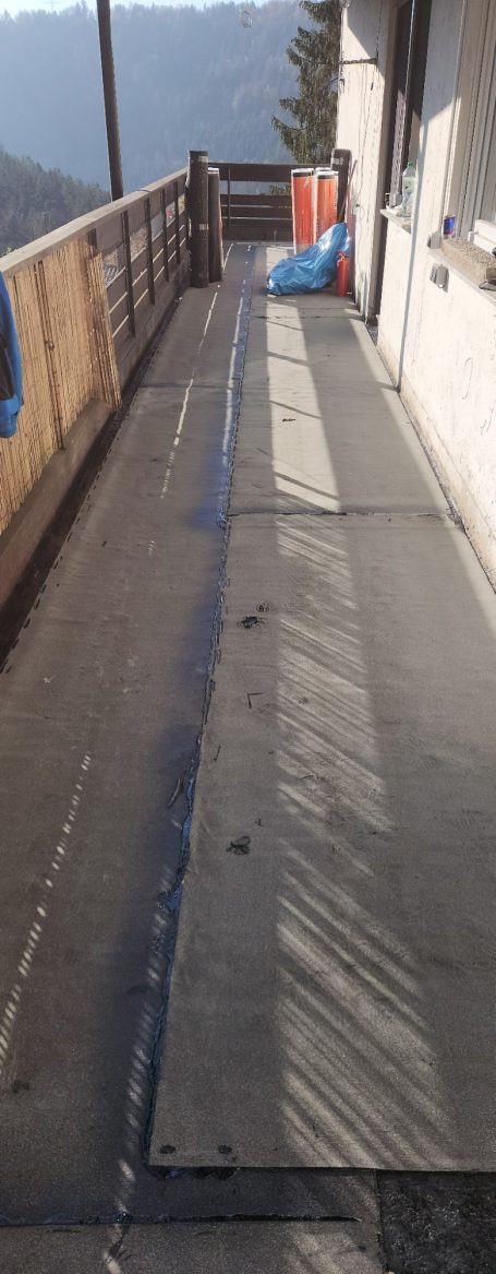 Balkon mit Betonfläche und Holzgeländer, im Hintergrund Berge und eine blaue Plane.